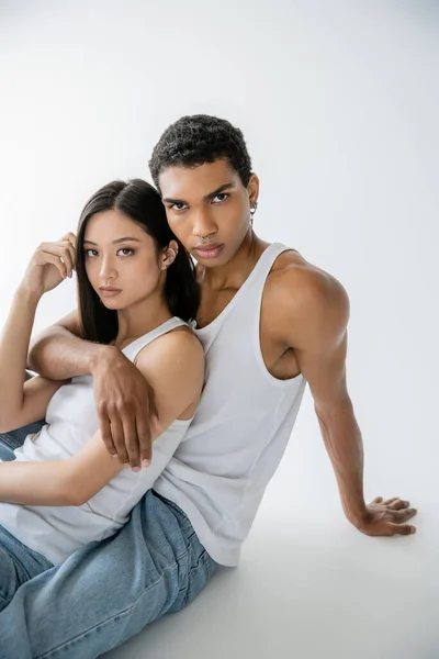 African american man in white tank top embracing sensual asian woman and looking at camera on grey background — Stock Photo