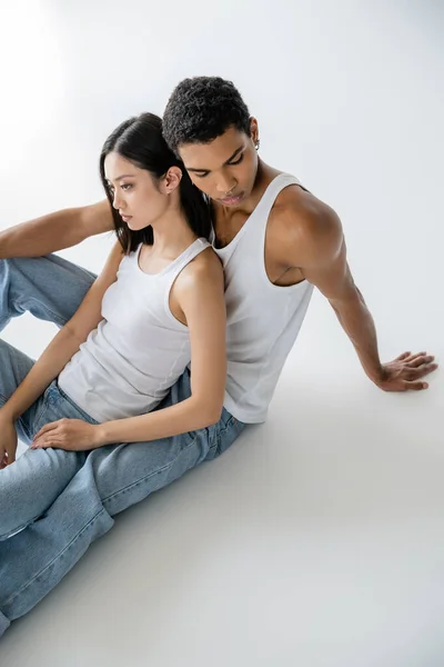 Высокий угол обзора молодых межрасовых пар в белых топах и джинсах, сидящих на сером фоне — стоковое фото