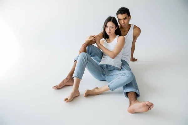 Longitud completa de la pareja multiétnica descalza sentado en pantalones vaqueros azules y camisetas blancas sobre fondo gris - foto de stock