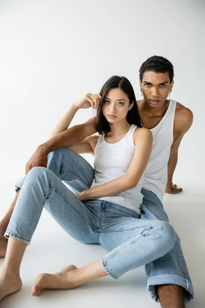 Modelos interracial con estilo en pantalones vaqueros azules y camisetas blancas sentados y mirando a la cámara sobre fondo gris - foto de stock