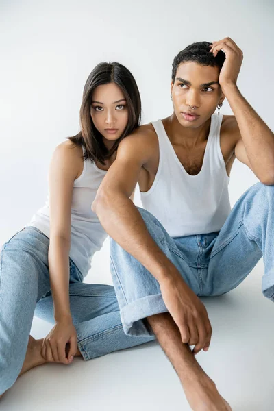 Brunette asian woman in tank top and jeans looking at camera near dreamy african american man sitting on grey background — Stock Photo