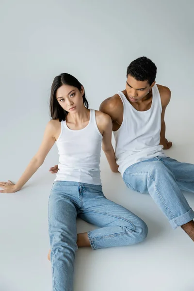 Slim asian woman in blue jeans and white tank top looking at camera near african american guy on grey background — Stock Photo