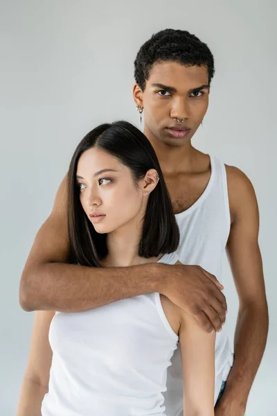 Afro-américain guy en blanc débardeur étreignant brunette asiatique modèle et regardant caméra isolé sur gris — Photo de stock
