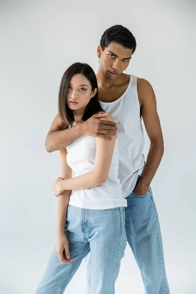 Young african american man holding hand in pocket of blue jeans and hugging asian woman isolated on grey — Stock Photo
