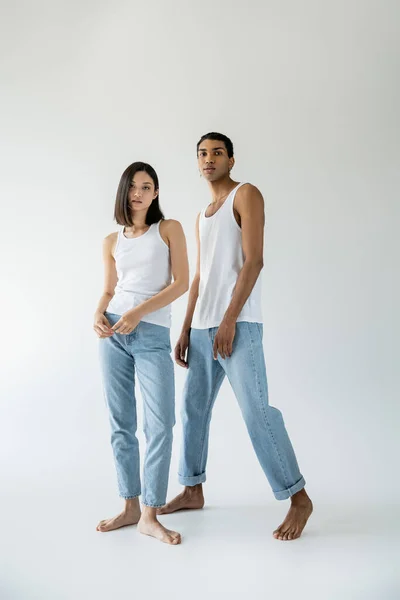 Longitud completa de la pareja interracial descalza y delgada en camisetas blancas y pantalones vaqueros posando sobre fondo gris - foto de stock