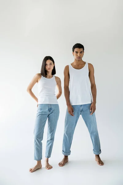 Полная длина молодых и босых мультиэтнических пар, позирующих в белых топах и синих джинсах на сером фоне — стоковое фото