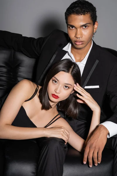 Joven y elegante pareja interracial posando en el sofá de cuero negro y mirando a la cámara sobre fondo gris - foto de stock