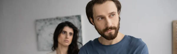 Dissatisfied bearded man looking away near blurred girlfriend at home, banner — Stock Photo