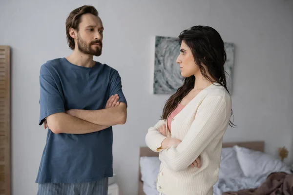 Femme offensée croisant les bras et regardant petit ami barbu en pyjama dans la chambre — Photo de stock