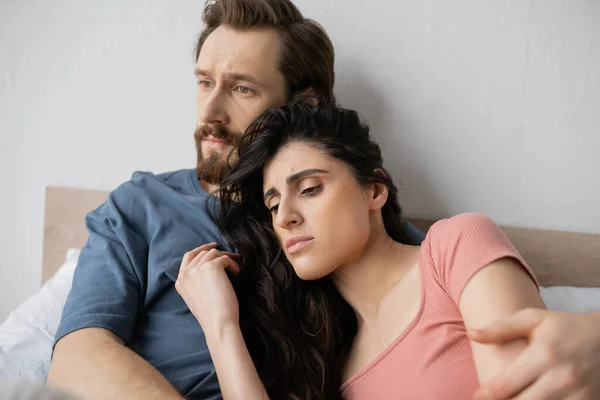 Triste uomo abbracciare bruna fidanzata in pigiama sul letto a casa — Foto stock