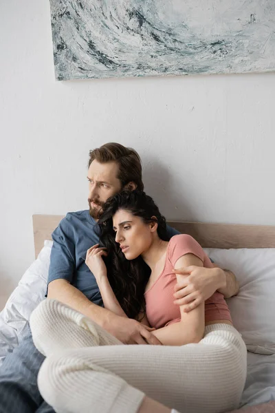Displeased man hugging brunette girlfriend in pajama on bed at home — Stock Photo