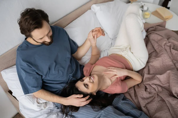 Top view of bearded man in pajama touching hair of cheerful girlfriend on bed at home — Stock Photo
