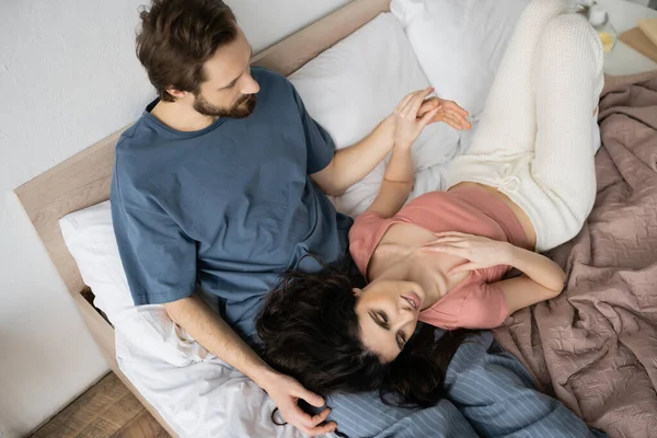 Draufsicht eines bärtigen Mannes, der die Hand einer lächelnden brünetten Freundin im heimischen Bett berührt — Stockfoto