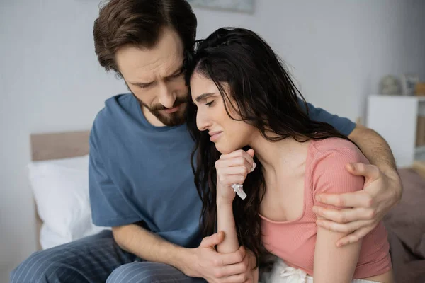 Hombre empático en pijama calmando a su novia llorando con servilleta en el dormitorio - foto de stock