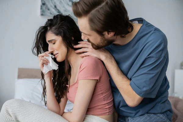 Uomo premuroso in pigiama abbracciare piangendo fidanzata con tovagliolo in camera da letto — Foto stock