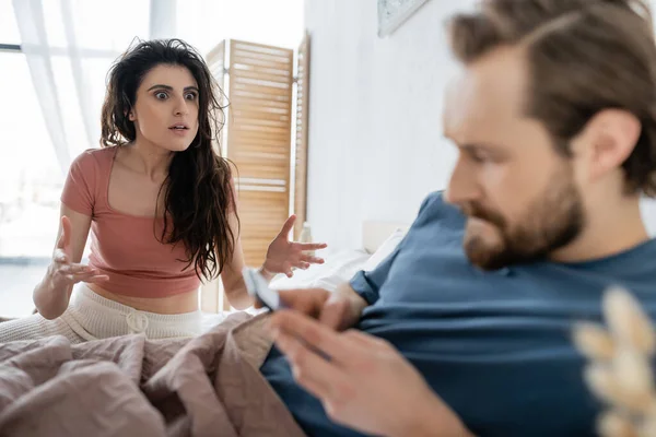 Annoyed woman in pajama quarrelling at blurred boyfriend with smartphone on bed — Stock Photo