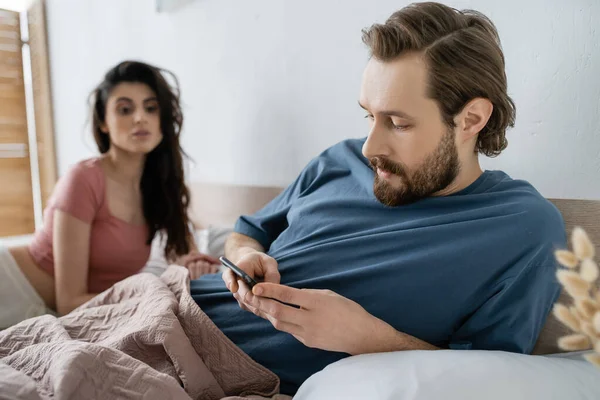 Бородатый мужчина, использующий мобильный телефон рядом с размытой девушкой в постели дома — стоковое фото