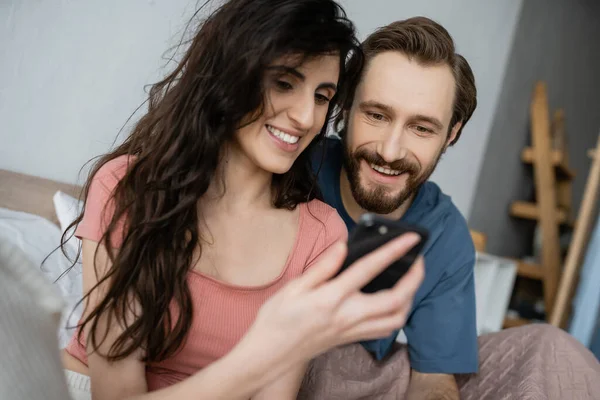 Усміхнена брюнетка жінка використовує розмитий смартфон біля хлопця на ліжку вдома — стокове фото