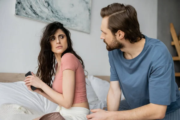 Mujer ofendida en pijama escondiendo teléfono inteligente cerca de novio celoso en el dormitorio - foto de stock