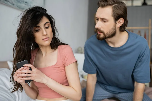 Morena mujer ocultando el teléfono celular cerca de novio celoso en el dormitorio - foto de stock