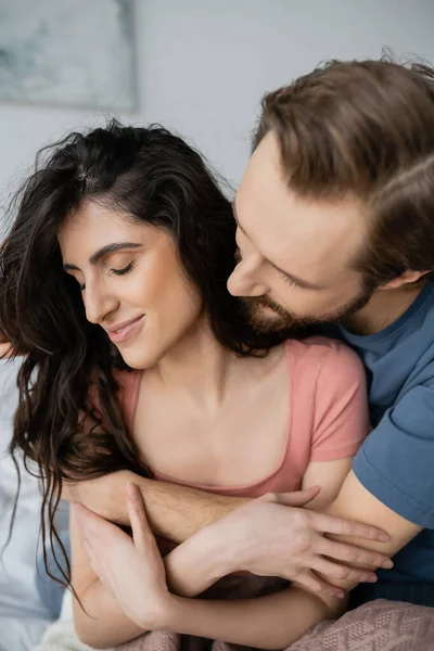 Bärtiger Mann umarmt lächelnde brünette Freundin im Schlafzimmer — Stockfoto