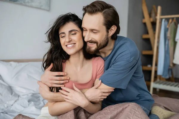 Exultante homem abraçando morena namorada no pijama na cama em casa — Fotografia de Stock