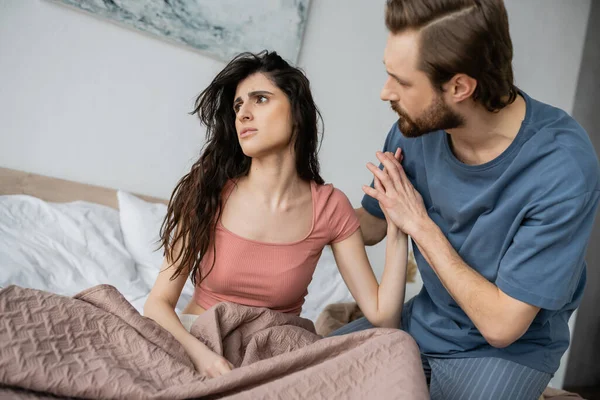Брюнетка мужчина трогает руку расстроенной и асексуальной девушки на кровати дома — стоковое фото