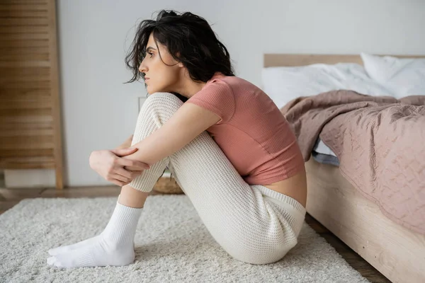 Vista laterale della donna depressa in pigiama seduta vicino al letto a casa — Foto stock