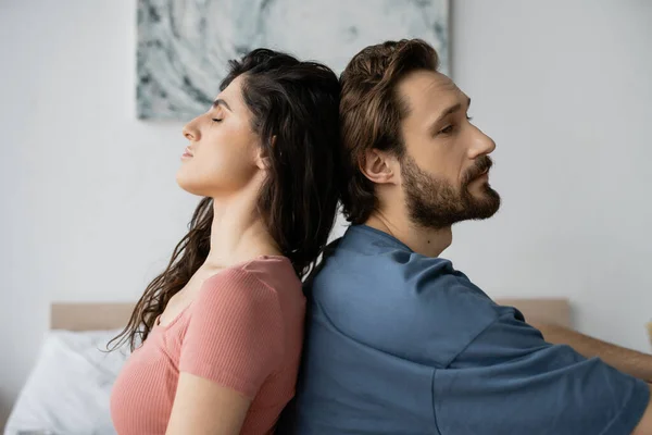 Upset bearded man sitting back to back with brunette girlfriend in bedroom — Stock Photo