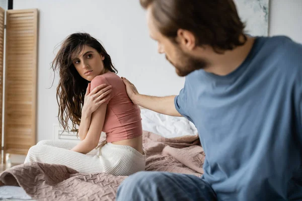 Размытый мужчина трогает обиженную девушку в пижаме на кровати дома — стоковое фото