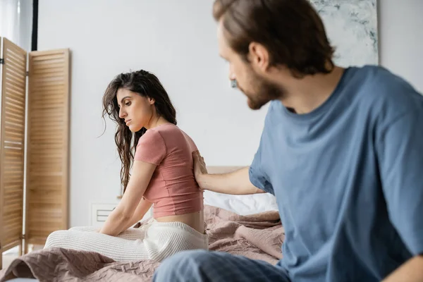 Blurred man touching disappointed girlified in pajama on bed — Stock Photo
