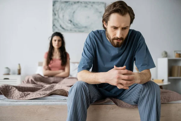 Uomo teso in pigiama seduto vicino alla fidanzata offuscata in camera da letto a casa — Foto stock