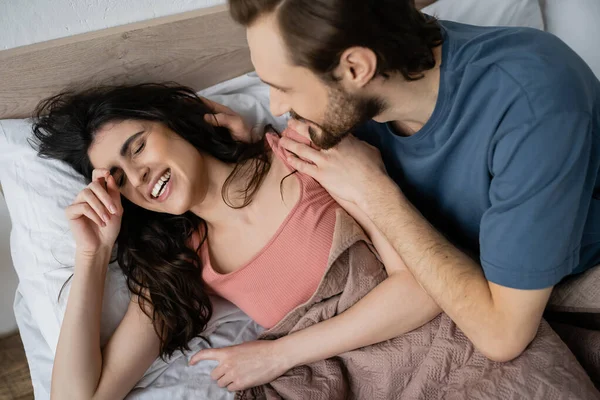 Vue grand angle de l'homme étreignant joyeuse petite amie brune sur le lit le matin — Photo de stock