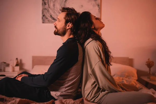 Vista lateral do casal com mal-entendido no relacionamento sentado de volta para trás na cama à noite — Fotografia de Stock