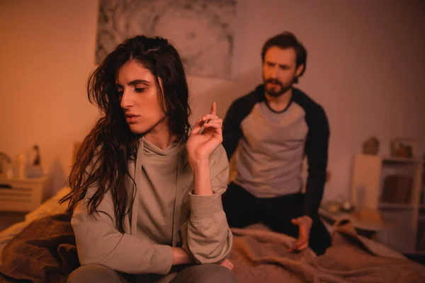 Sad brunette woman sitting near blurred boyfriend on bed at night — Stock Photo