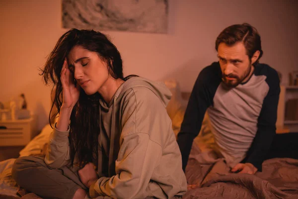 Расстроенная женщина, сидящая рядом с расплывчатым бойфрендом и разговаривающая по вечерам в постели — стоковое фото