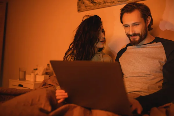 Femme brune parlant à petit ami souriant près d'un ordinateur portable flou sur le lit la nuit — Photo de stock