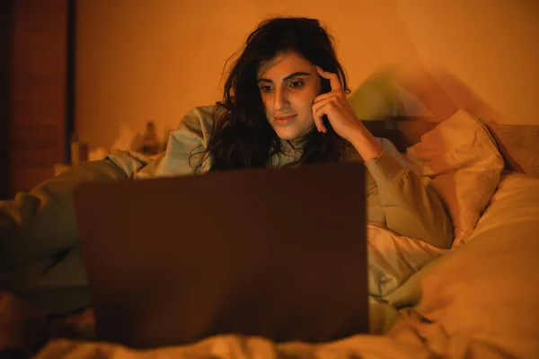 Mulher morena olhando para laptop desfocado na cama à noite — Fotografia de Stock