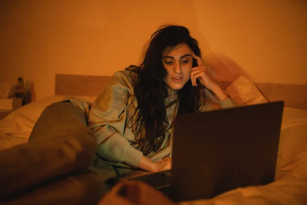 Morena mulher assistindo filme no laptop enquanto deitado na cama à noite — Fotografia de Stock