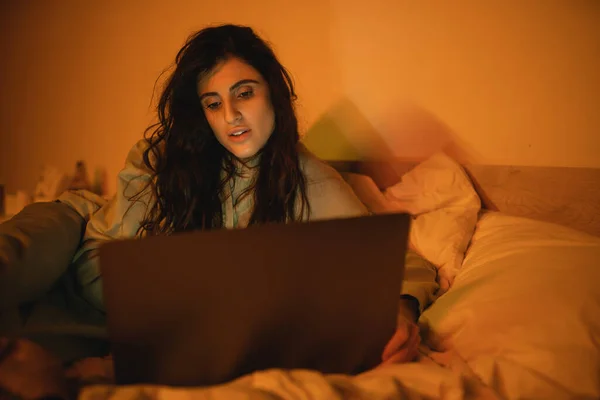 Brunette woman using blurred laptop while lying on bed at night — Stock Photo