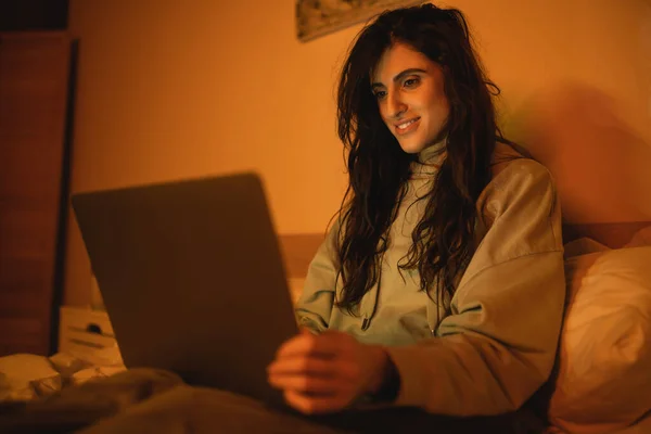 Lächelnde brünette Frau mit verschwommenem Laptop nachts auf dem Bett — Stockfoto