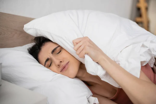 Mujer morena molesta poniendo almohada en la cabeza en el dormitorio por la mañana - foto de stock
