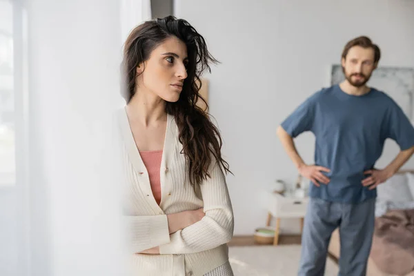 Angry woman crossing arms while standing near blurred boyfriend in bedroom — Stock Photo