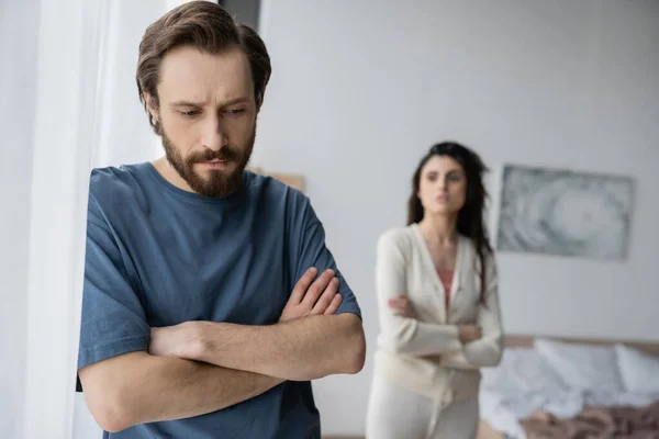 Offended man crossing arms while standing near curtain and blurred girlfriend at home — Stock Photo