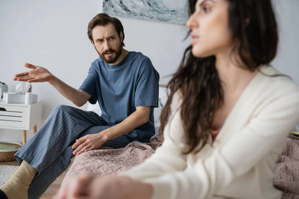 Обиженный мужчина в пижаме разговаривает с расплывчатой девушкой, сидя дома на кровати — стоковое фото