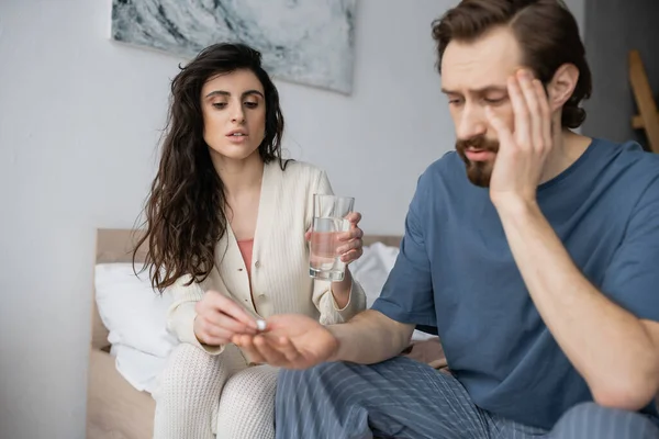 Caring woman giving pill and holding water near blurred boyfriend in bedroom at home — Stock Photo