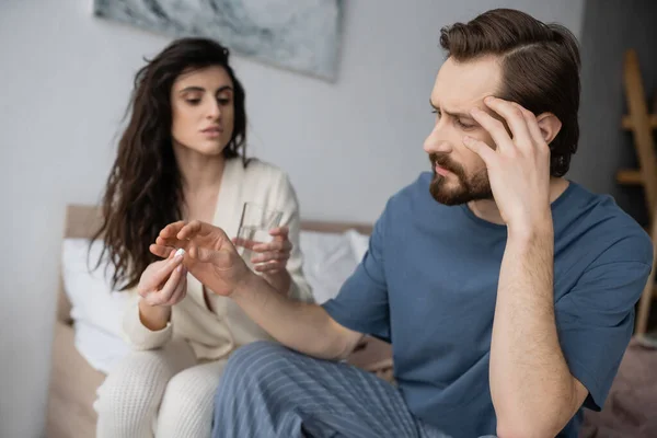 Man with headache taking pill from blurred caring girlfriend in bedroom — Stock Photo