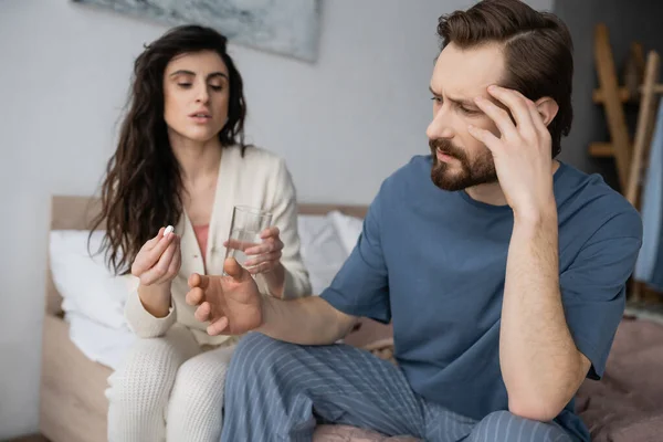 Bearded man with migraine sitting near blurred girlfriend with pills and water at home — Stock Photo