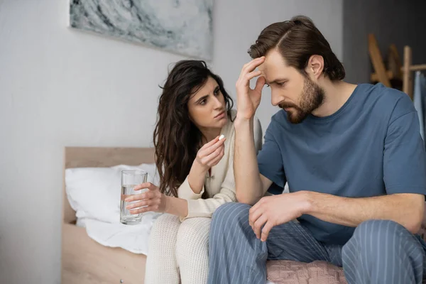 Заботливая женщина с таблетками и стаканом воды рядом с парнем, страдающим мигренью в спальне — стоковое фото