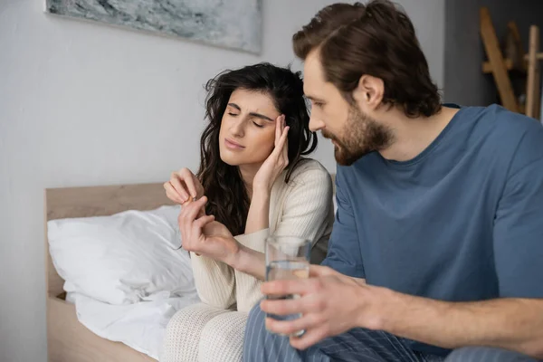 Заботливый мужчина дает таблетки и воду девушке с мигренью в спальне — стоковое фото